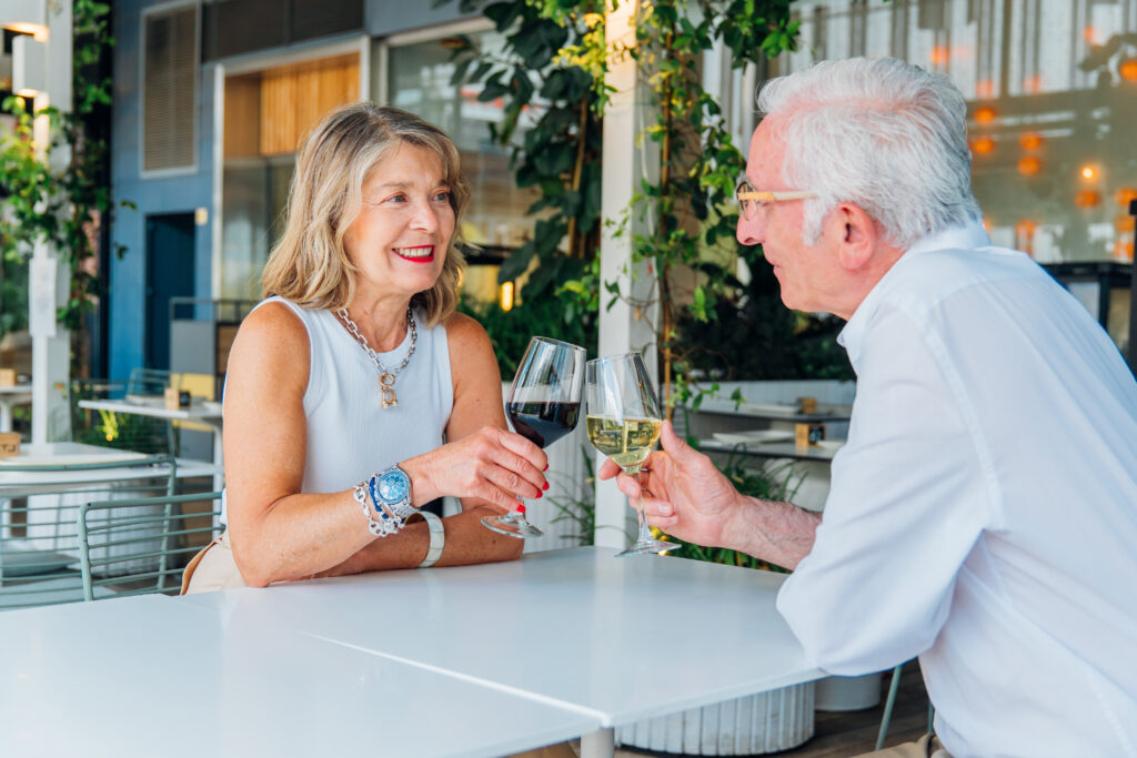 Happy elderly couple toasting with glasses of wine while sitting at a table in an outdoor restaurant, celebrating their union and enjoying a romantic moment.